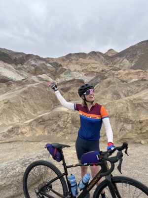 Caeli on Climate Ride RDV waving beside her bike before majestic sand dunes.